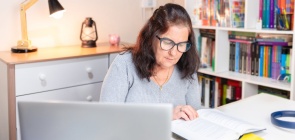 Professora estudando em casa. Ela está com o computador aberto e faz anotações em caderno. No fundo, uma estante de livros