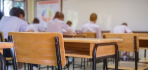 sala de aula, com alunos ao fundo. Em primeiro plano, carteiras vazias