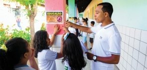 Professor conversa com alunas e aponta para cartaz