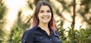 Mulher jovem está parada em frente a um fundo de natureza. Ela tem cabelos castanhos, pele clara e mexas cor-de-rosa no cabelo. Está vestindo uma blusa azul de botões.