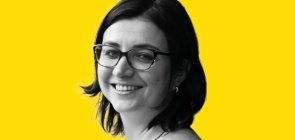 Mulher de óculos, está de lado e sorri para a foto. Ela tem cabelo na altura do ombro