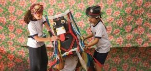 Em um fundo colorido, aparece uma menina usando um microfone e uniforme segurando um boi de papelão com fitas colorida. Também segurando o boi aparece uma menina com chapeu sertanejo. Dentro da caixa do boi, uma criança aparece