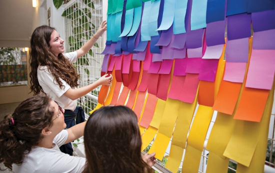 Professora mostra a alunos painel com pedaços de papel coloridos
