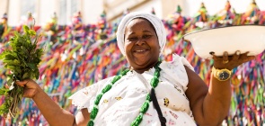 Foto colorida de Sacerdotisa do Candomblé, uma mulher negra, vestindo roupas tradicionais da religião, de cor branca, e cordões coloridos, em frente a igreja do Bonfim, em Salvador, Bahia, Brasil