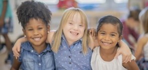 Três alunas, uma de inclusão ao centro, se abraçam e olham para a câmera sorrindo em uma sala de aula