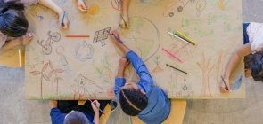 Um grupo de cinco crianças pequenas vistas de cima desenhando em torno de um papel pardo