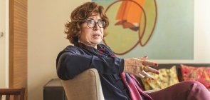 Mulher branca com cabelo curto cor mel, usa óculos, está sentada em uma poltrona conversando