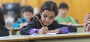 aluna concentrada escrevendo em um caderno, dentro de uma sala de aula.