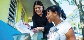 Em primeiro plano uma mulher com um computador sorri e olha para onde aponta mulher em pé
