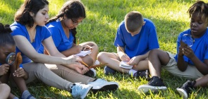 estudantes, sentados na grama, fazem anotações em cadernos. Um deles segura uma lupa e observa uma folha