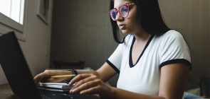 Fotografia de aluna do ensino médio em frente ao computador fazendo anotações em seu caderno.