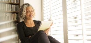 Uma mulher lê um livro sorrindo. Ela tem cabelos grisalhos cacheados e usa roupa preta. 