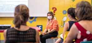 Uma mulher loira de camiseta laranja e calça pretaestá sentada à frente de outras três mulheres, que estão de costas. Todas assistem a uma palestra em uma escola. Ao fundo, uma parede amarela com pinturas de símbolos como pontos de interrogação e aspas