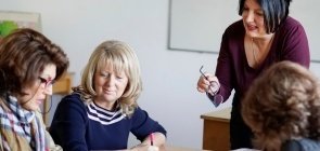 Três professoras de cerca de 40 anos sentadas juntas em uma mesa com uma quarta mulher de pé olhando as atividades