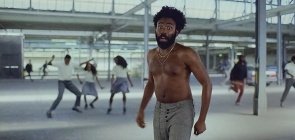 Trecho do videoclipe This is America do Childish Gambino. Na foto aparece ele no meio, homem negro ao redor dos 30 anos, sem camisa, usa uma calça social cinza. No fundo jovens com uniformes dançam
