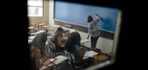 Professora dá aula para alunos em sala de aula. Todos de costas, não é possível identificar.