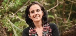 Mulher sentada em troco de árvore sorri para a foto. No fundo há galhos e folhas