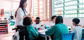 Estudantes reunidos em grupo, eles estão sentados e a professora está de pé
