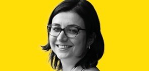 Mulher de óculos, está de lado e sorri para a foto. Ela tem cabelo na altura do ombro