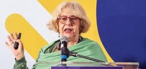 Senhora loira vestindo um casaco verde e um xale verde, usa óculos discursa 