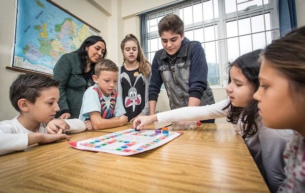 Seis alunos em volta de uma mesa olham concentrados para um tabuleiro. Uma menina mexe uma peça. Uma professora acompanha a atividade