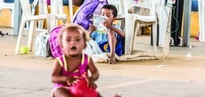 Uma bebê em primeiro plano aparece embaçada com um brinquedo na mão. No fundo, um menino sentado no chão, apoiado numa cadeira desgastada, lê