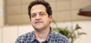 Homem branco de cabelo escuro e curto olha para a foto. Ele veste camisa xadrez e segura um lápis