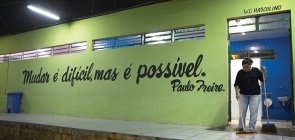 Em uma parede verde lê-se mudar é dificil, mas é possível de Paulo Freire. Ao lado, uma mulher limpa o banheiro masculino