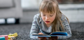 Foto de uma menina de 8 anos com sindrome de down, ela lê deitada no chão 