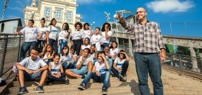 Homem de aproximadamente 30 anos aponta e alunos, sentados olham, no fundo vê-se a igreja da Penha