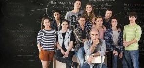 Um grupo de dez adolescentes, sentado em destaque tem um senhor mais velho, sentado e olhando de maneira séria para a câmara. No fundo, uma lousa com conteúdo de filosofia