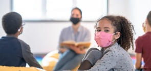 Uma menina usa máscara cor de rosa e olha para a câmera enquanto duas pessoas aparecem desfocadas ao fundo