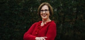 Senhora de 70 anos veste casaco vermelho, ela sorri. No fundo um espaço arborizado