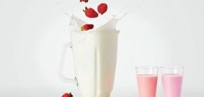 Num fundo branco, aparece no centro um liquidificador com um líquido branco, caindo aparecem morangos que espalham o líquido. Ao lado dois copos, um com um líquido rosa escuro e outro mais claro
