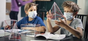 Dois meninos de máscara estudam sobre triângulos. Um deles segura um triângulo transparente enquanto a professora aparece ao fundo, desfocada. 