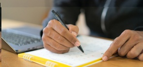 Foto de uma pessoa escrevendo, na mesa um computador e um calendário