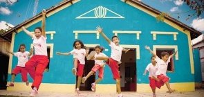 Foto com crianças pulando. Elas vestem um uniforme de short vermelho e camiseta branca. No fundo uma casa azul