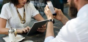 Sem mostrar o rosto, aparecem um homem e uma mulher. Ela de camiseta branca e colar marrom mexe em um tablet e ele de camisa social branca mexe no celular. Na mesa onde estão sentados está um chá gelado e um açucareiro