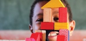 Uma criança sorrindo, na sua frente há uma casa feita de blocos coloridos de madeira