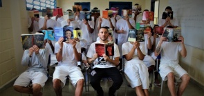 Foto colorida de professor em sala de aula, diante de sala cheia de alunos. Os alunos estão de costa para a imagem e veste camisas brancas. Ao fundo, tela de televisão, mesa de professor e paredes cor bege.