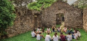 Em um sítio arqueológico, um grupo de alunos senta em roda no gramado