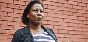 Mulher negra tem cabelo curto preto, usa vestido listrado e uma jaqueta