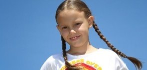Ao ar livre, menina branca de cabelo comprido, loiro e com duas trancinhas, sorri para a foto e veste uma camiseta branca com um arco-iris