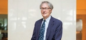 Homem branco sorri, tem cabelo grisalho, usa óculos, veste terno azul com camisa social azul claro e gravata azul com estampa. Usa sapato social preto, ele está encostado em poltrona marrom. 