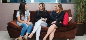 Sentadas em um sofá preto, três mulheres conversam