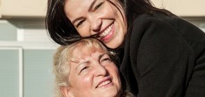 Mulher branca de 36 anos, tem cabelo preto liso, veste um casaco preto. Ela sorri e abraçoa uma senhora loira de casaco branco e jaleco preto