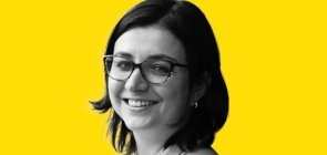 Mulher de óculos, está de lado e sorri para a foto. Ela tem cabelo na altura do ombro