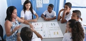 Em uma mesa, seis alunos olham para a professora que está na ponta e apresenta um jogo de tabuleiro que está no centro da mesa