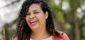 Mulher negra de cabelo cacheado, blusa branca e casaco rosa sorri para a foto sem olhar