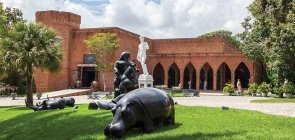Foto do Instituto Brennand, uma área verde com monumentos e ao fundo uma estrutura de tijolos